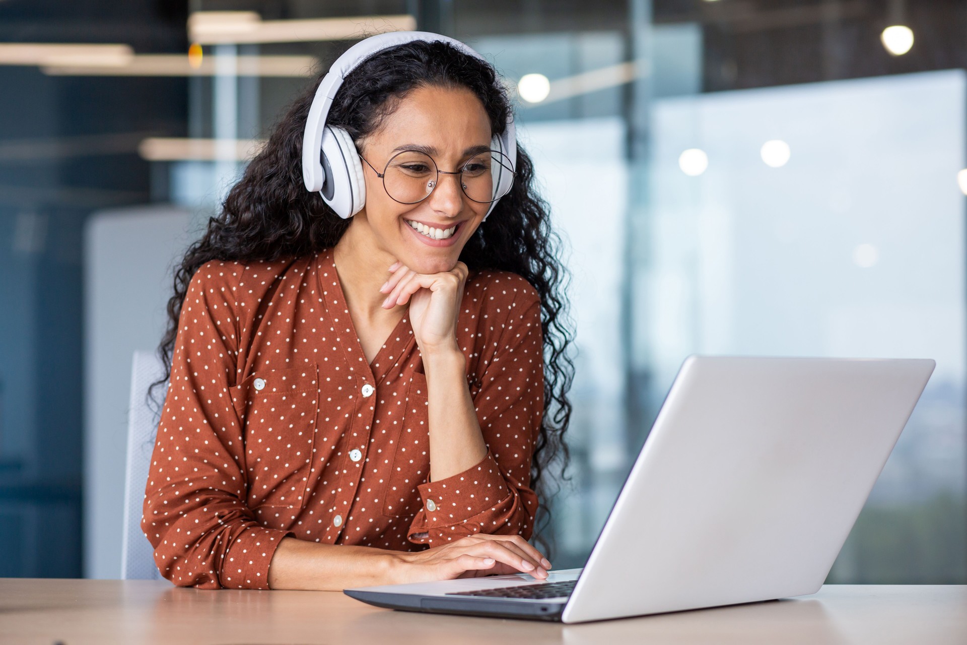 Young beautiful woman in headphones working with laptop inside office at workplace, joyful and satisfied business woman watching online webinar, educational video course, learning remotely